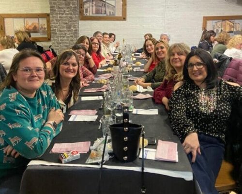 En la imagen se ve a un grupo de mujeres sonrientes sentadas a una mesa de restaurante de cena.